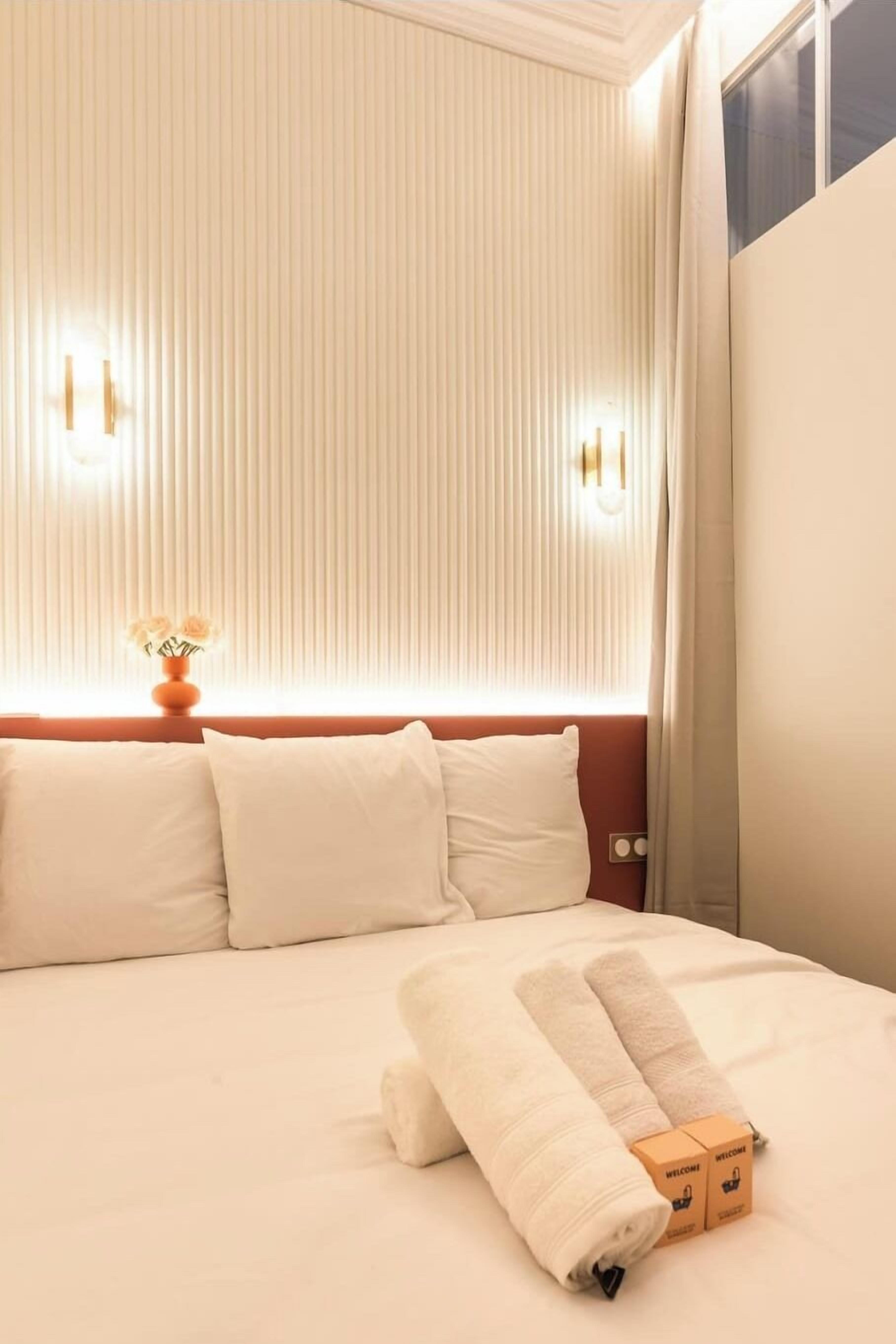 Una cama bien hecha con una toalla doblada y dos cajas de bienvenida, un florero con flores, y una lámpara fijada a la pared.