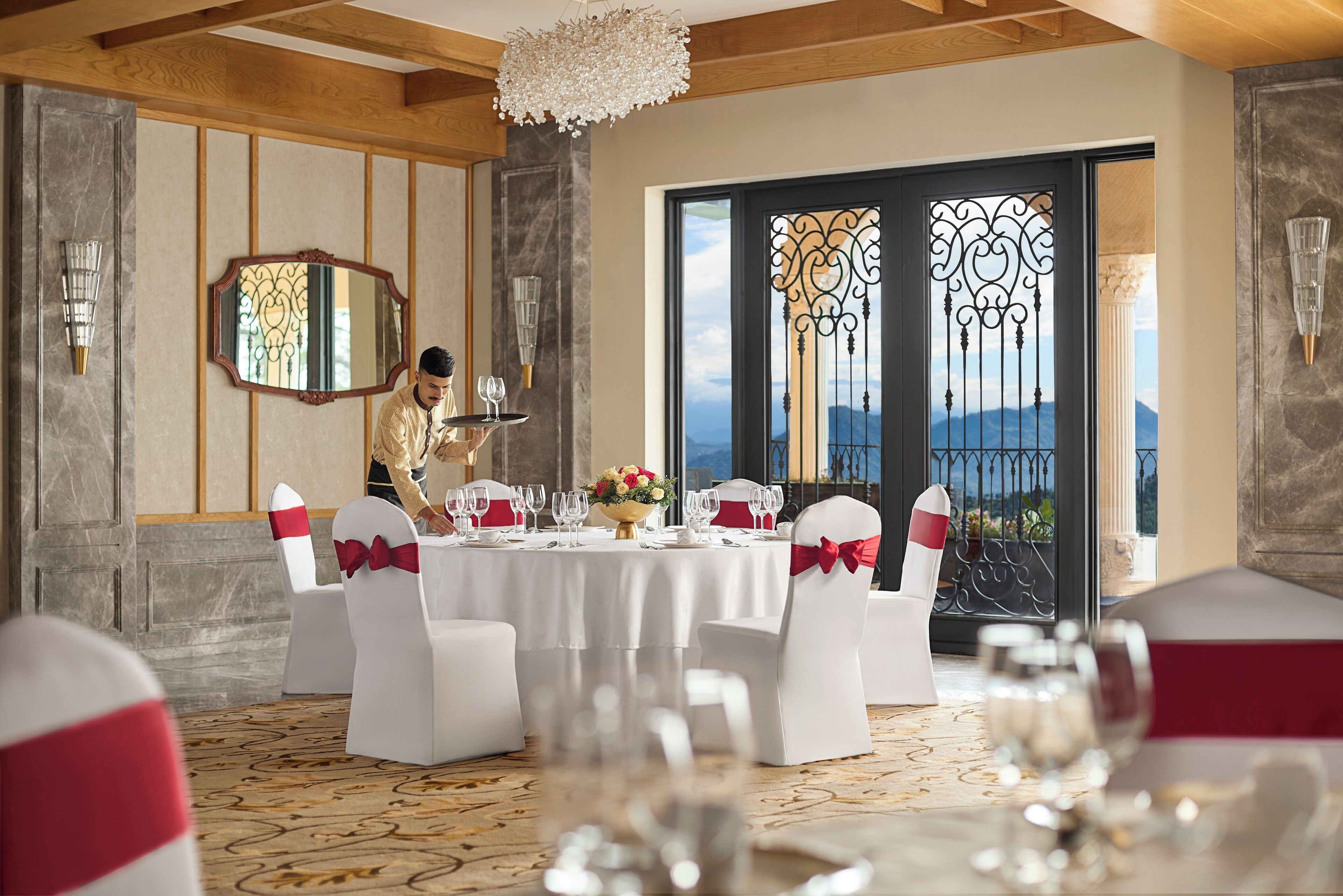 Un comedor formal con una mesa dispuesta para una comida, sillas blancas con cintas rojas y un espejo decorativo en la pared.