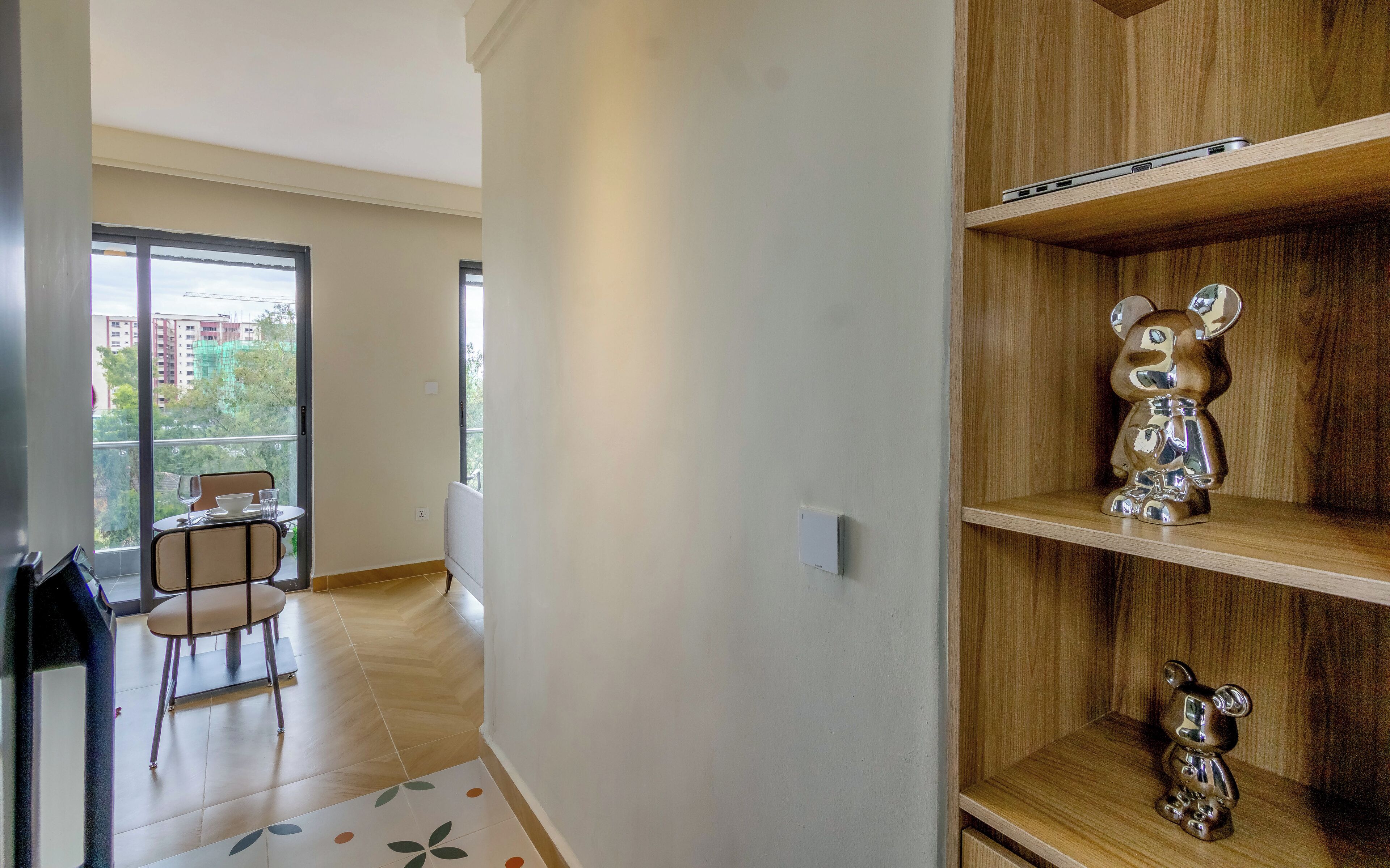 Un apartamento moderno con comedor, una estantería de madera con adornos y vistas a edificios a través de amplios ventanales.