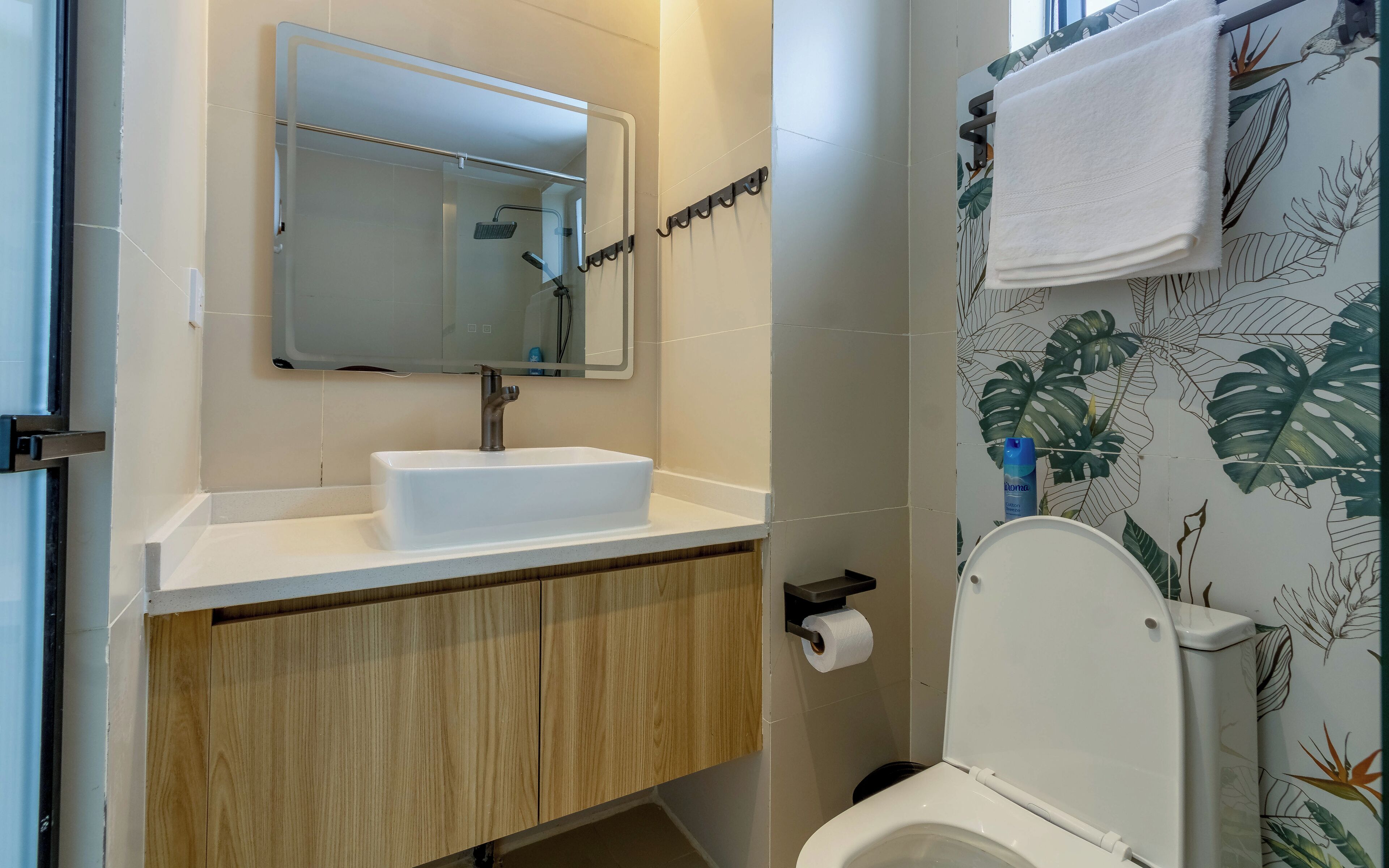 Ein Badezimmer mit einem weißen Waschbecken, einem Spiegel und einer Toilette. Über dem Waschbecken befindet sich ein Handtuchhalter und an der Wand ein Toilettenpapierhalter.