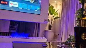 A modern living room with a flat-screen TV, a small table with a laptop, and a potted plant.