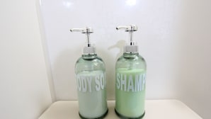 Zwei Glasflaschen mit Pumpspendern, beschriftet mit 'BODY SOAP' und 'SHAMPOO', auf einer weißen Oberfläche.