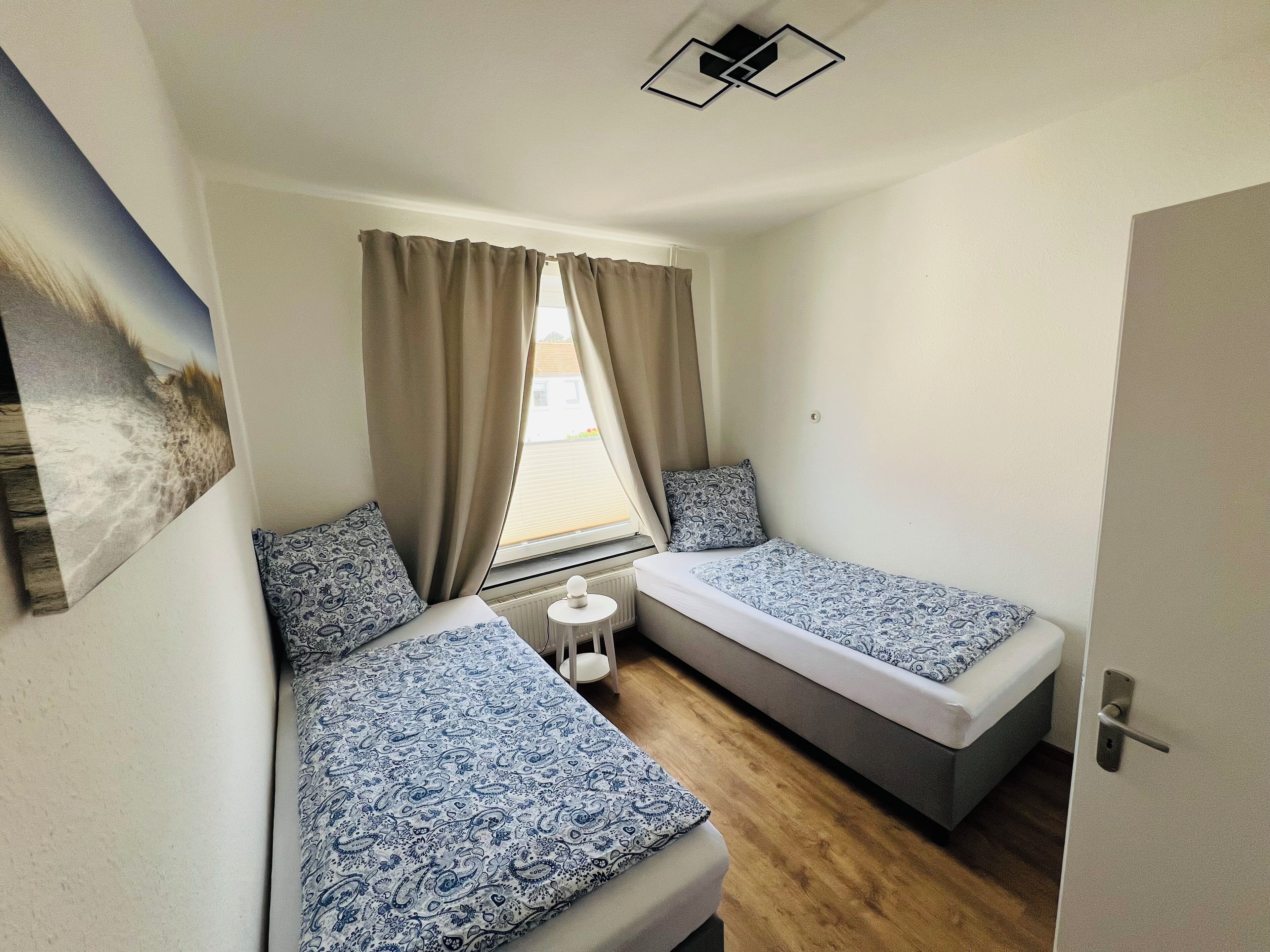 Quarto pequeno e compacto com duas camas de solteiro, uma mesinha e uma janela com cortinas.