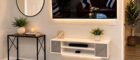 Una sala de estar con un televisor de pared, una mesa consola con adornos, una planta en maceta y un espejo en la pared.