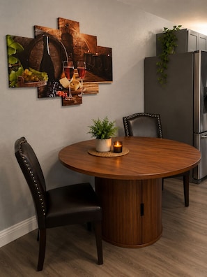 A dining area with a round wooden table, two upholstered chairs, a wall-mounted wine rack, and a stainless steel refrigerator.