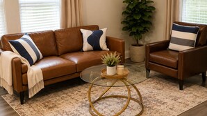 A living room with a brown leather sofa, a brown leather armchair, a glass coffee table, a potted plant, and a ceiling fan.