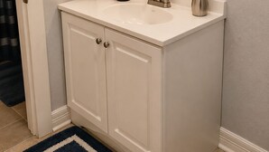 A bathroom with a white vanity, a mirror with lights, a sink, a faucet, a soap dispenser, and a small plant.