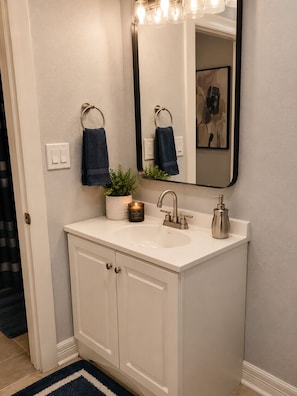 A bathroom with a white vanity, a mirror with lights, a sink, a faucet, a soap dispenser, and a small plant.