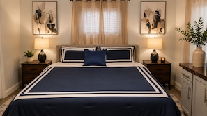 A large bed with a navy blue and white bedspread, flanked by two bedside tables with lamps, a potted plant, and artwork on the walls.