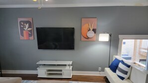 A living room with a flat-screen TV mounted on the wall, a white entertainment unit below it, a beige sofa, a patterned rug, and a window with a view of a wooden deck.