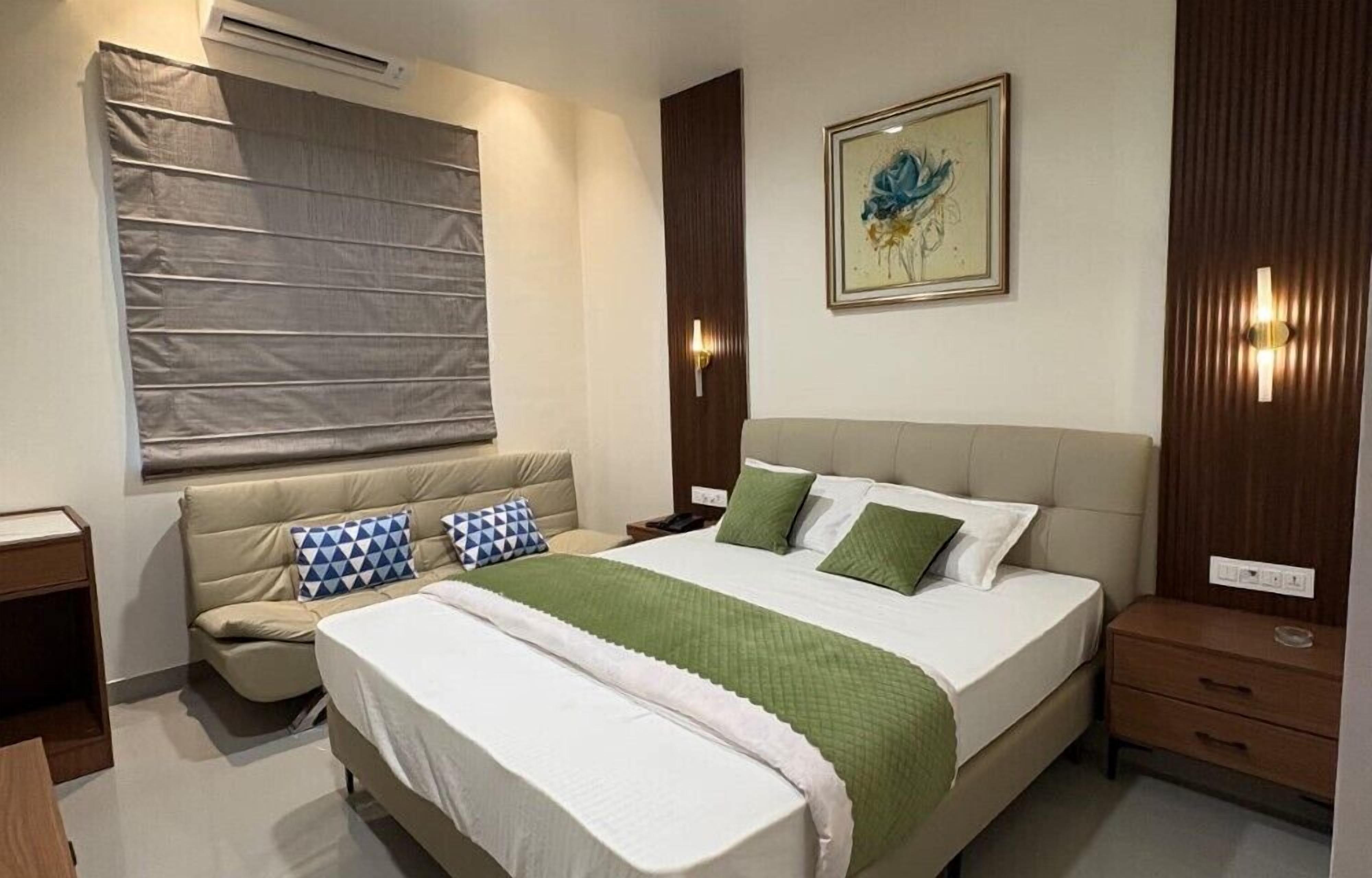 A bedroom with a bed, bedside tables, a nightstand, a wall-mounted air conditioner, a window with blinds, and a framed picture on the wall.