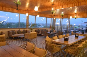 An indoor seating area with tables and chairs, large windows, and hanging plants. - Hotel Mountain Paradise (Abu Road)