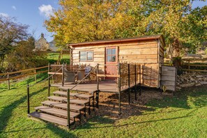 A rustic wooden cabin with a porch, steps, and a fenced area. - Private Cotswold Shepherd’s Hut with Garden & Countryside Views (Charlton)