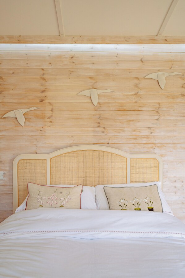 Una cama con cabecera trenzada, dos almohadas con bordado floral y paneles de madera con siluetas de pájaros.