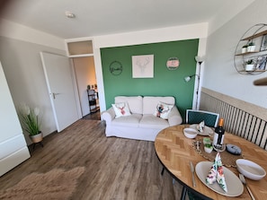 A living room with a white sofa, a dining table set with plates and a bottle, a green accent wall, and a clock. - Apartment Schwarzwald Heimat with Terrace, Pool, Sauna, and Fitness (Schönwald)