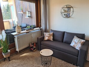 Un salon chaleureux avec un canapé foncé, une table basse ronde, un radiateur, une fenêtre avec des rideaux et une étagère murale agrémentée de plantes.