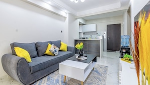 A modern living room with a grey sofa, a white coffee table, and a kitchen in the background. - Yaya Suites (Nairobi)