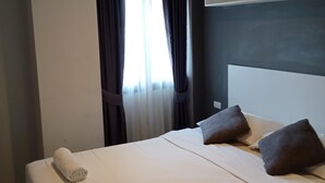 A hotel room with a large bed, two pillows, a rolled towel, a window with curtains, and a shelf above the bed. - Well-located condo 1-BR T1-NV 203 (Chalong)