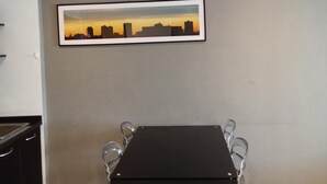 A dining area with a black table and chairs, a framed cityscape picture on the wall, and a kitchen in the background. - Well-located condo 1-BR T1-NV 203 (Chalong)