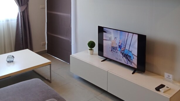 A modern living room with a flat-screen TV on a white entertainment unit, a small potted plant, and a sofa. - Well-located condo 1-BR T1-NV 203 (Chalong)