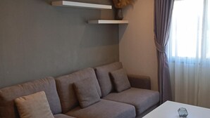 A modern living room with a sectional sofa, a small table, a mounted air conditioner, and a window with curtains. - Well-located condo 1-BR T1-NV 203 (Chalong)