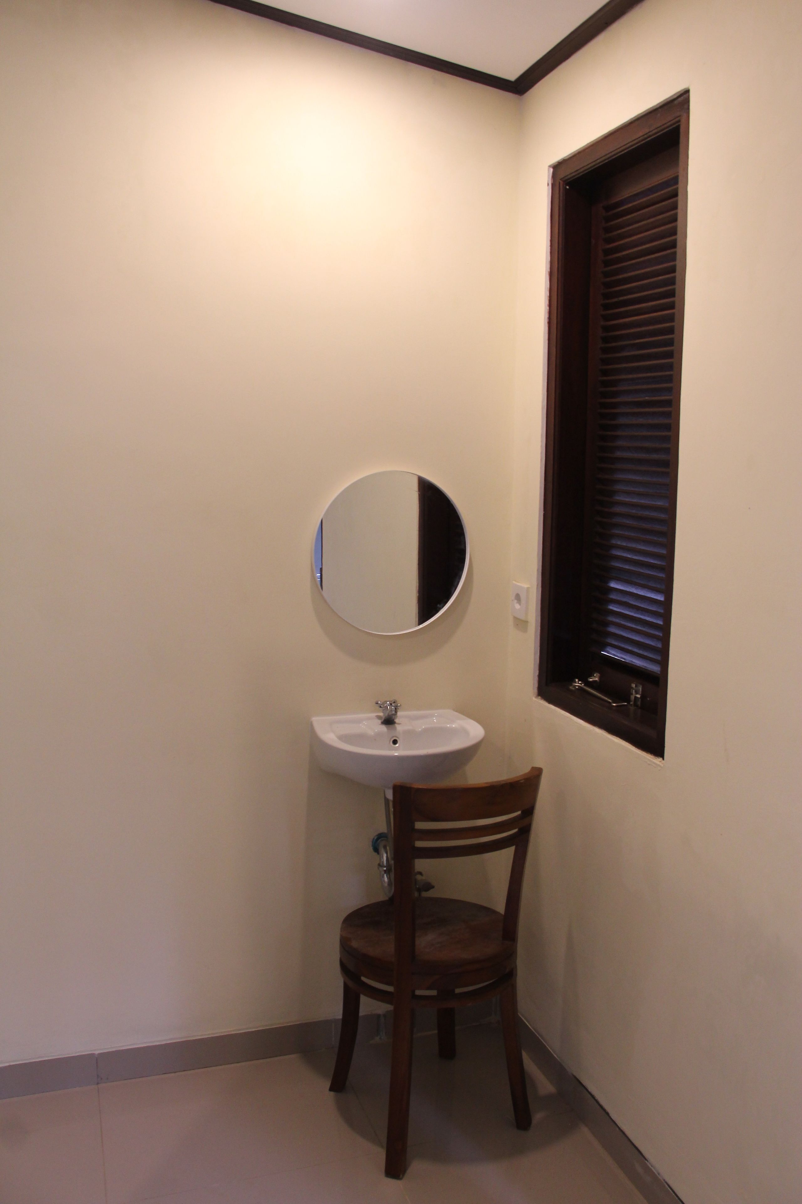 Un baño pequeño con un lavamanos blanco, una silla de madera y una ventana con persianas.