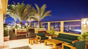 Terraço no telhado com móveis de madeira, almofadas verdes e plantas em vasos, com vista para a cidade à noite.