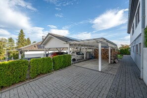 A covered parking area with a car and a truck, a paved driveway, and a building with a white facade.