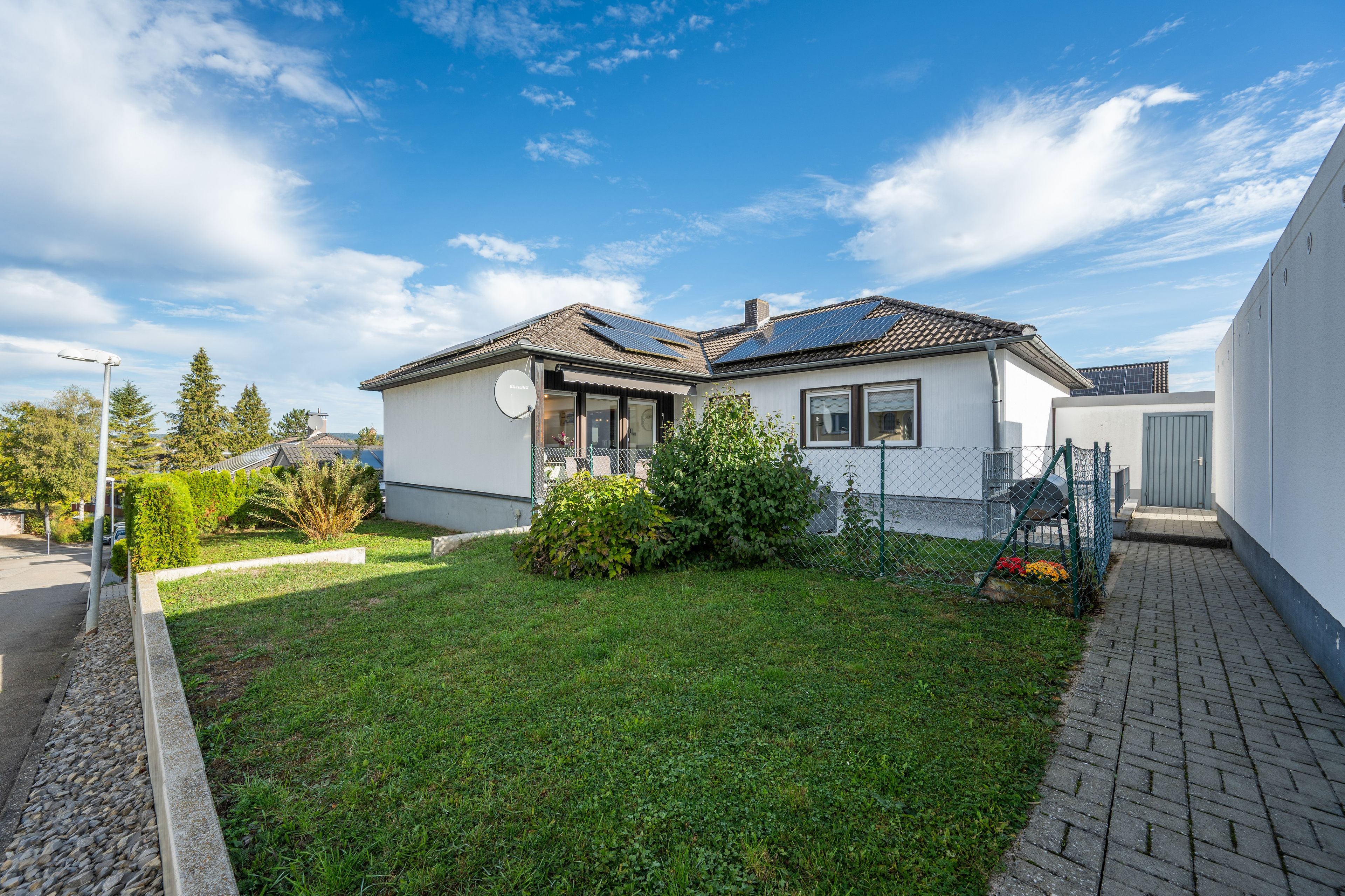 Ein Wohnhaus mit Solaranlage auf dem Dach, einem umzäunten Garten und einer gepflasterten Auffahrt.