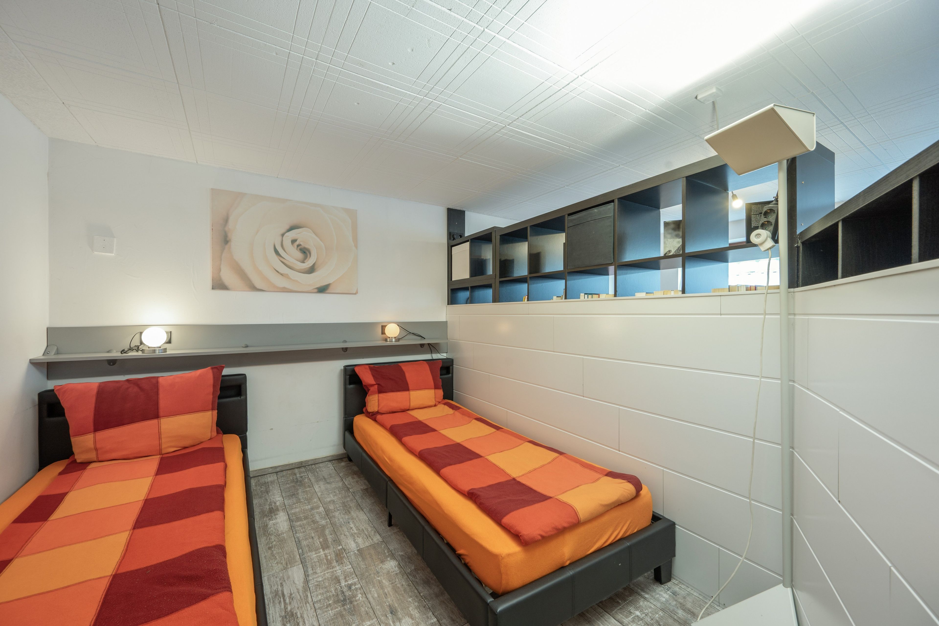 Ein Zimmer mit zwei Einzelbetten, einem an der Wand befestigten Bücherregal, einer Deckenleuchte und einer Steckdose.
