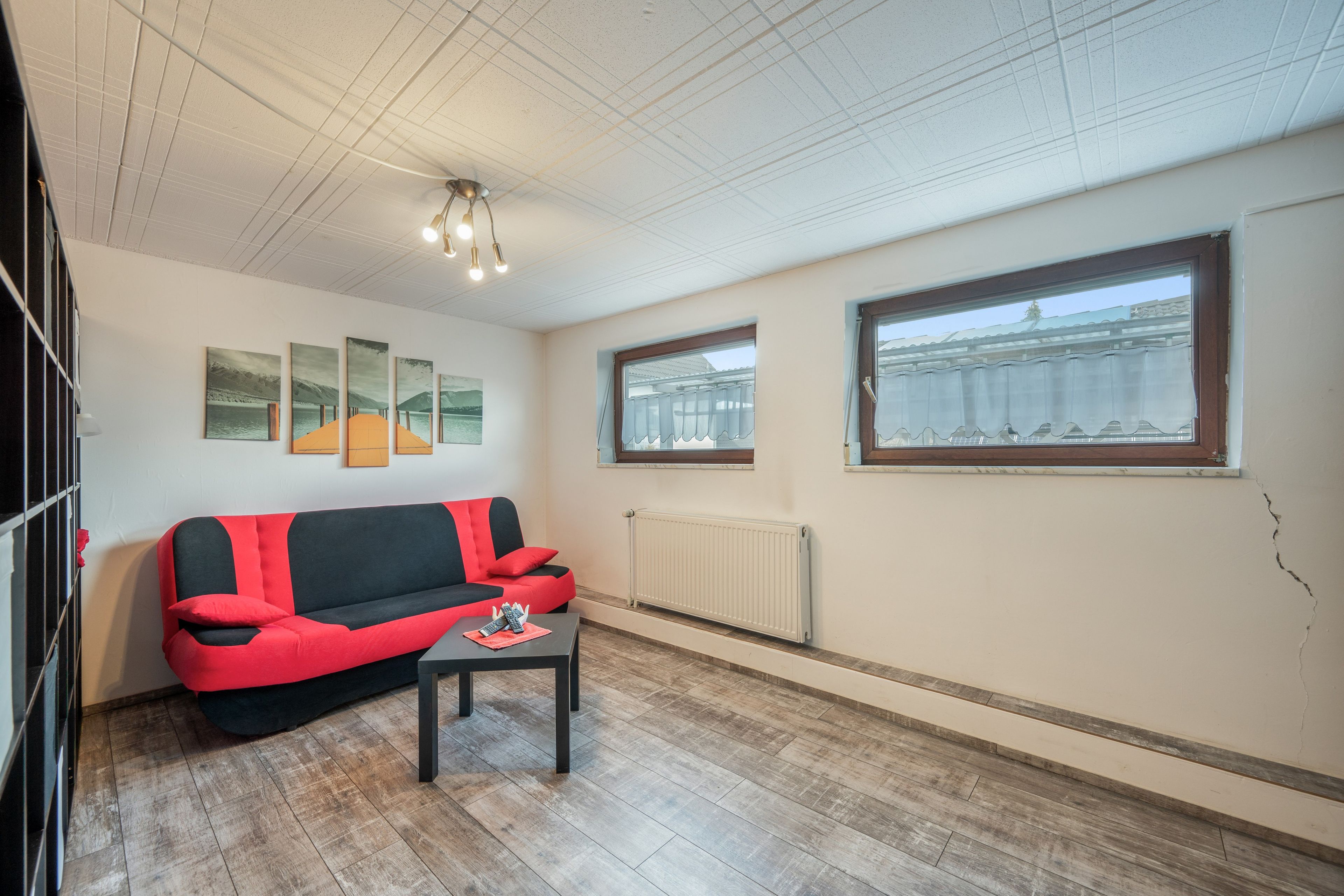 A room with a red and black sofa, a small table with remotes, a radiator, and a window with a view of a building.