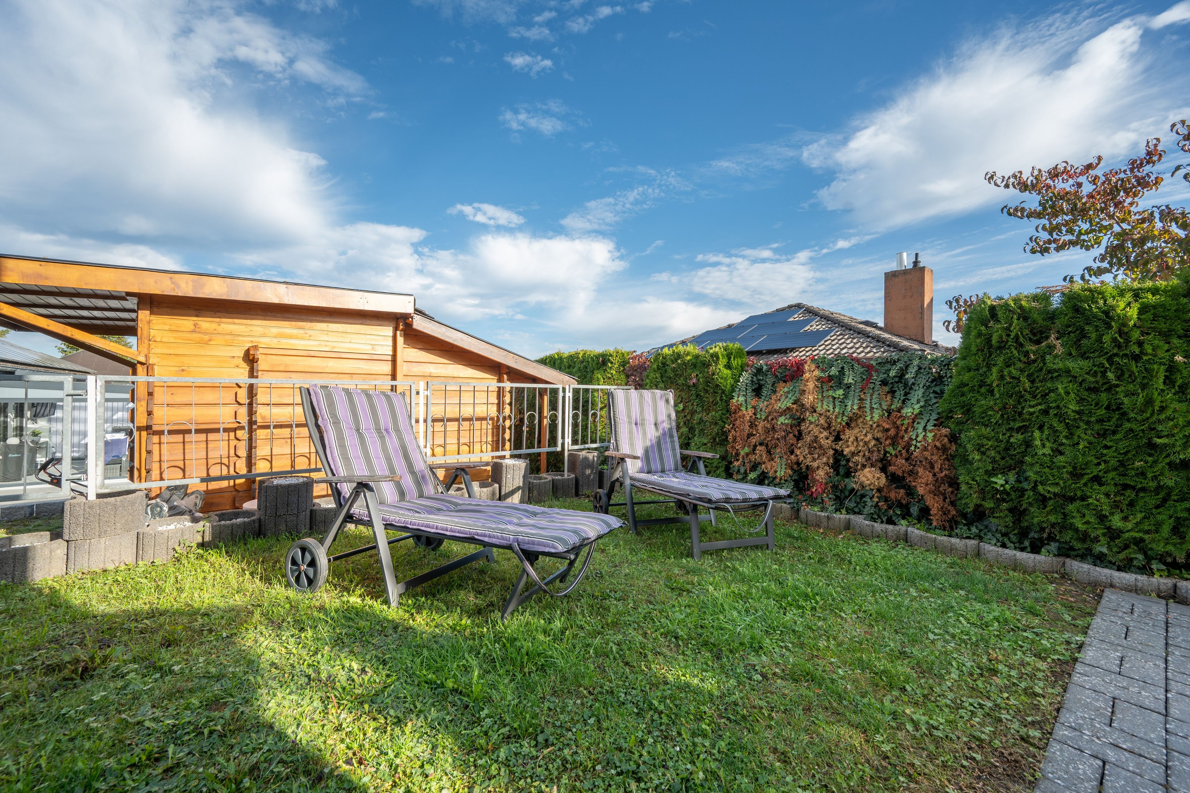 A wooden house with a garden, two sun loungers, and a metal fence.