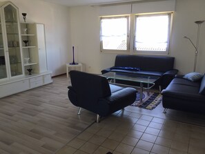 Uma sala de estar com sofá, mesa de centro de vidro, aparador com portas de vidro e janela com persianas.