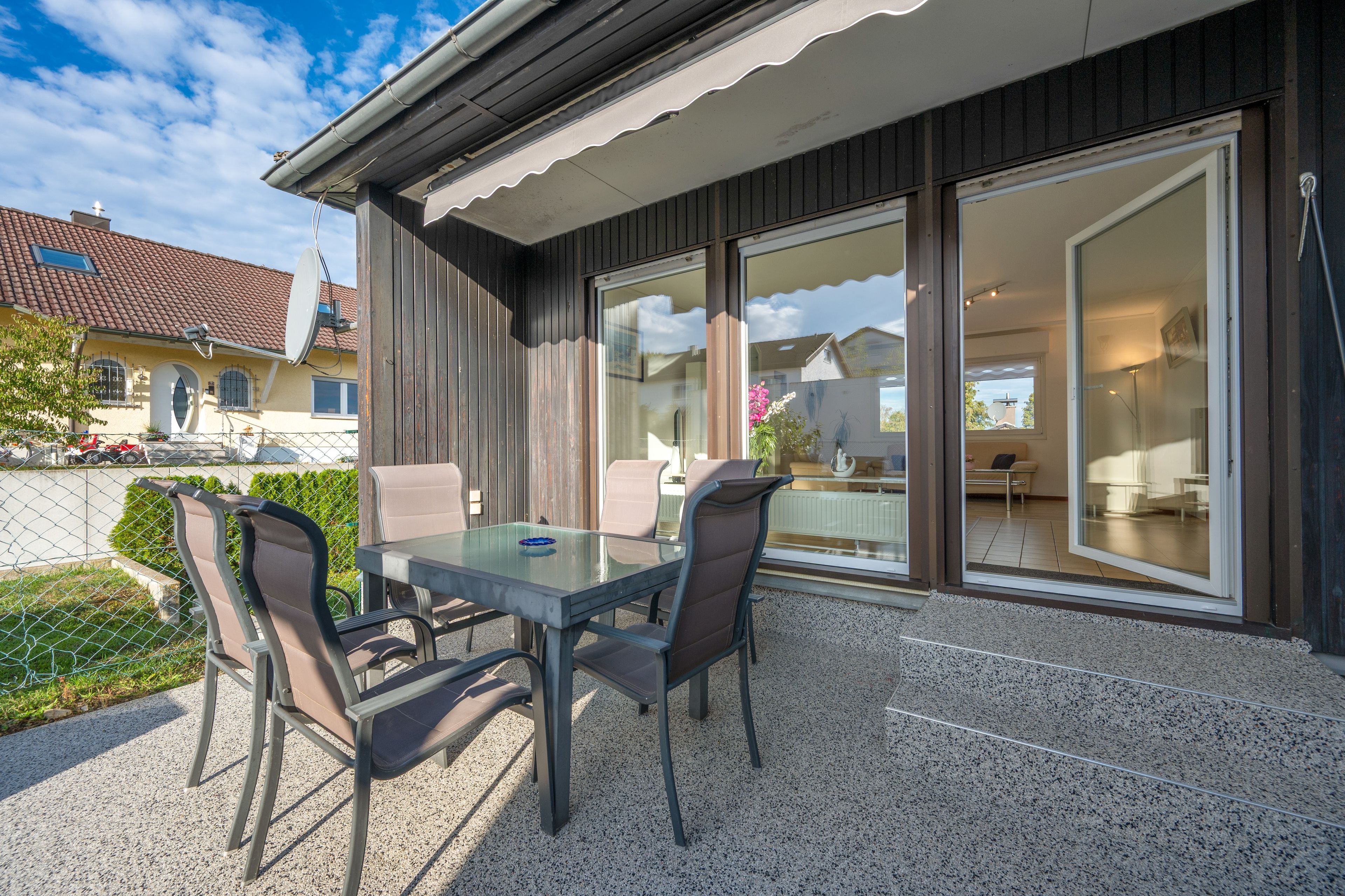 Een terras buiten met een glazen tafel en stoelen, een houten hek en uitzicht op een woongebouw.
