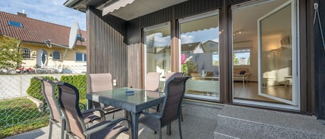 En udendørs terrasse med et glasbord og stole, et træhegn og udsigt til et boligkompleks.