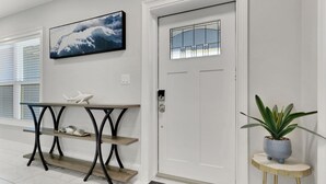 Een nette, moderne entree met een witte deur, een klein bijzettafeltje met een plant, een aan de muur bevestigd kunstwerk en een console tafel met decoratieve objecten.