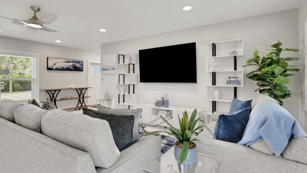 Een moderne woonkamer met een grijze bank, een salontafel, een aan de muur bevestigde televisie en een plafondventilator.
