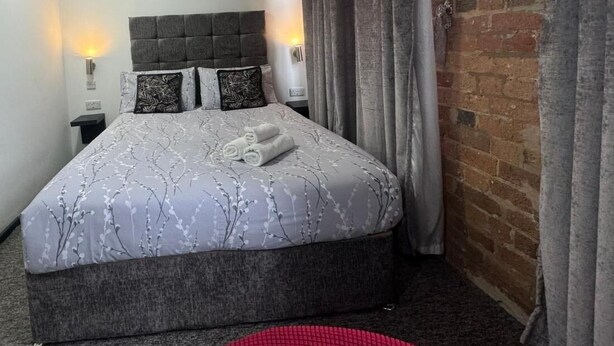 Un lit bien fait, avec une tête de lit grise et une literie à motifs, un tapis rond rouge et un mur en briques.