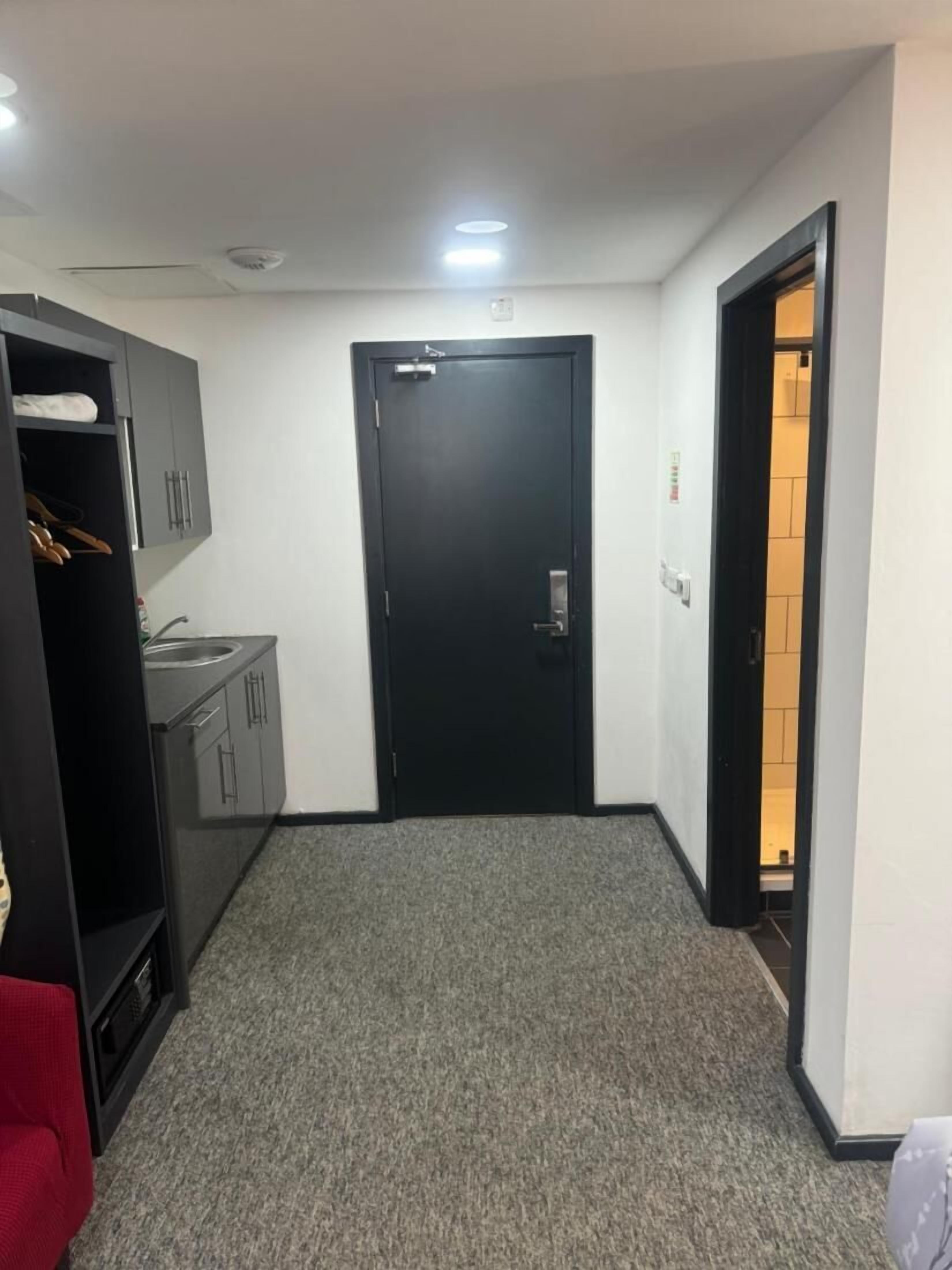 A small, clean room with a black door, a wardrobe, a small kitchenette, and a red chair.