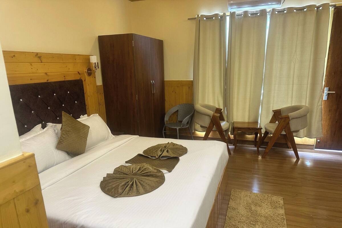 A hotel room with a bed, wooden headboard, wardrobe, two chairs, a small table, and a window with curtains.