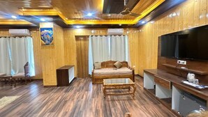 A room with a wooden floor, a sofa, a coffee table, a TV, and a ceiling fan. - The Chail Pine Orchard Resort (Chail)