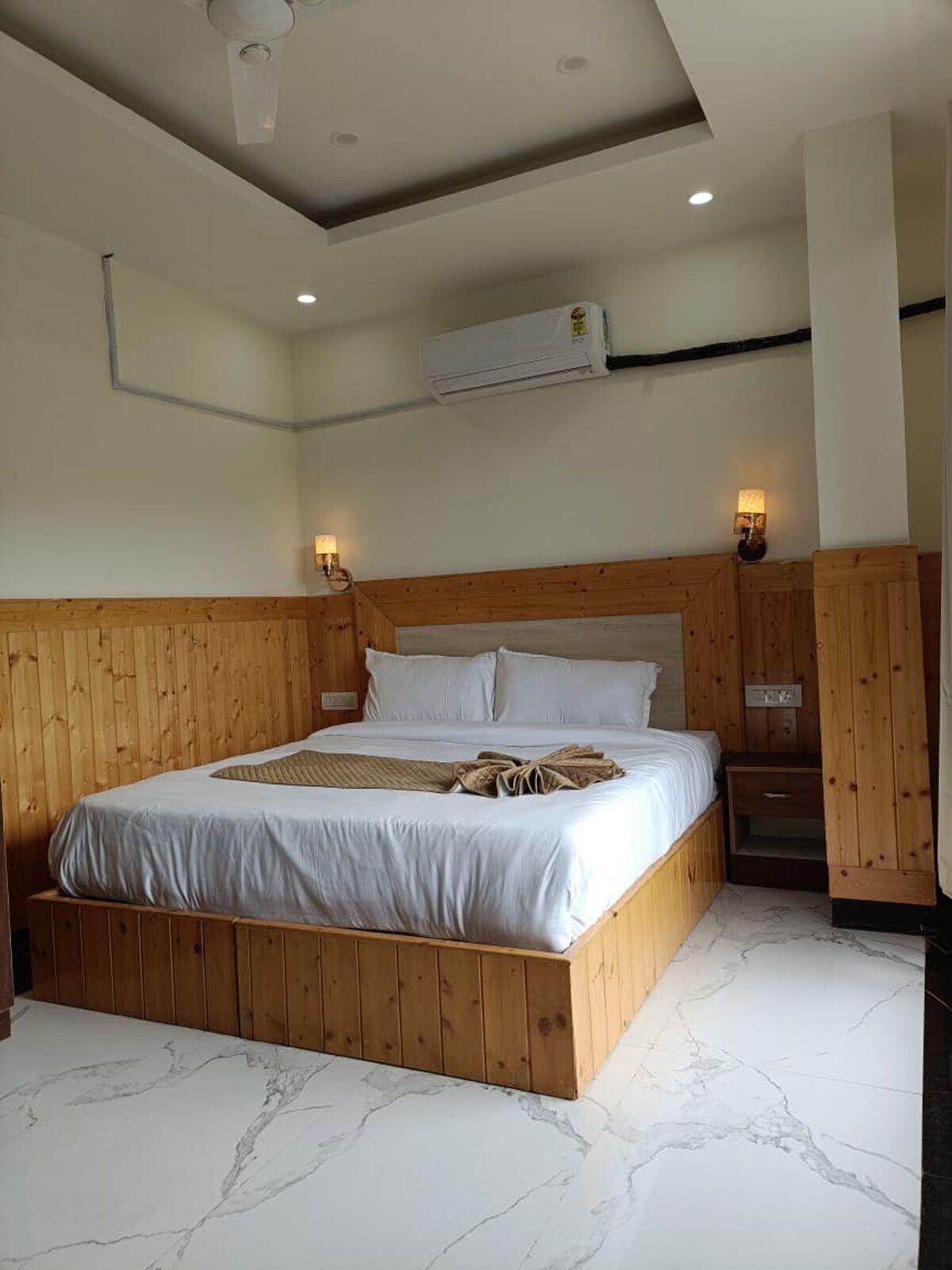 A bedroom with a wooden bed frame, white bedding, marble flooring, and two wall-mounted lamps.