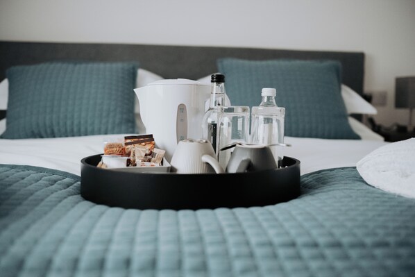 A neatly arranged tray on a bed with a white kettle, bottles, and a cup. The bed has a blue quilt and two pillows.