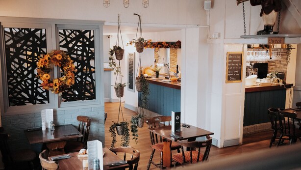A restaurant interior with wooden tables and chairs, hanging plants, and a kitchen area visible through a partition. - Fox and Hounds Inn (Carmarthen)