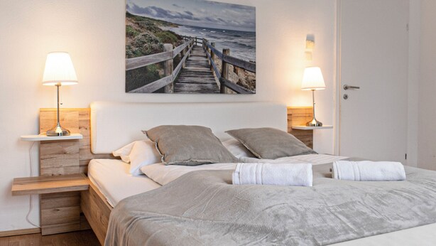 A bedroom with a wooden headboard, two bedside tables with lamps, a large bed with grey bedding, and a picture of a coastal walkway on the wall. - Bright 2BR Apt Perfect Long Stay (Vienna)