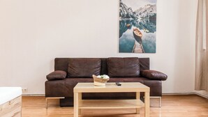A living room with a brown leather sofa, a wooden coffee table with a basket of flowers, and a painting of a mountainous lake on the wall. - Bright 2BR Apt Perfect Long Stay (Vienna)