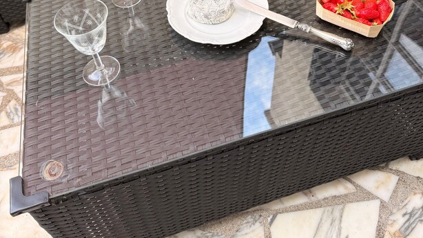 A wicker sofa with cushions, a glass table with a plate, a bottle, and a bowl of strawberries. - Gîte Du Centre with Private Terrace and Wi-Fi (Romorantin-Lanthenay)