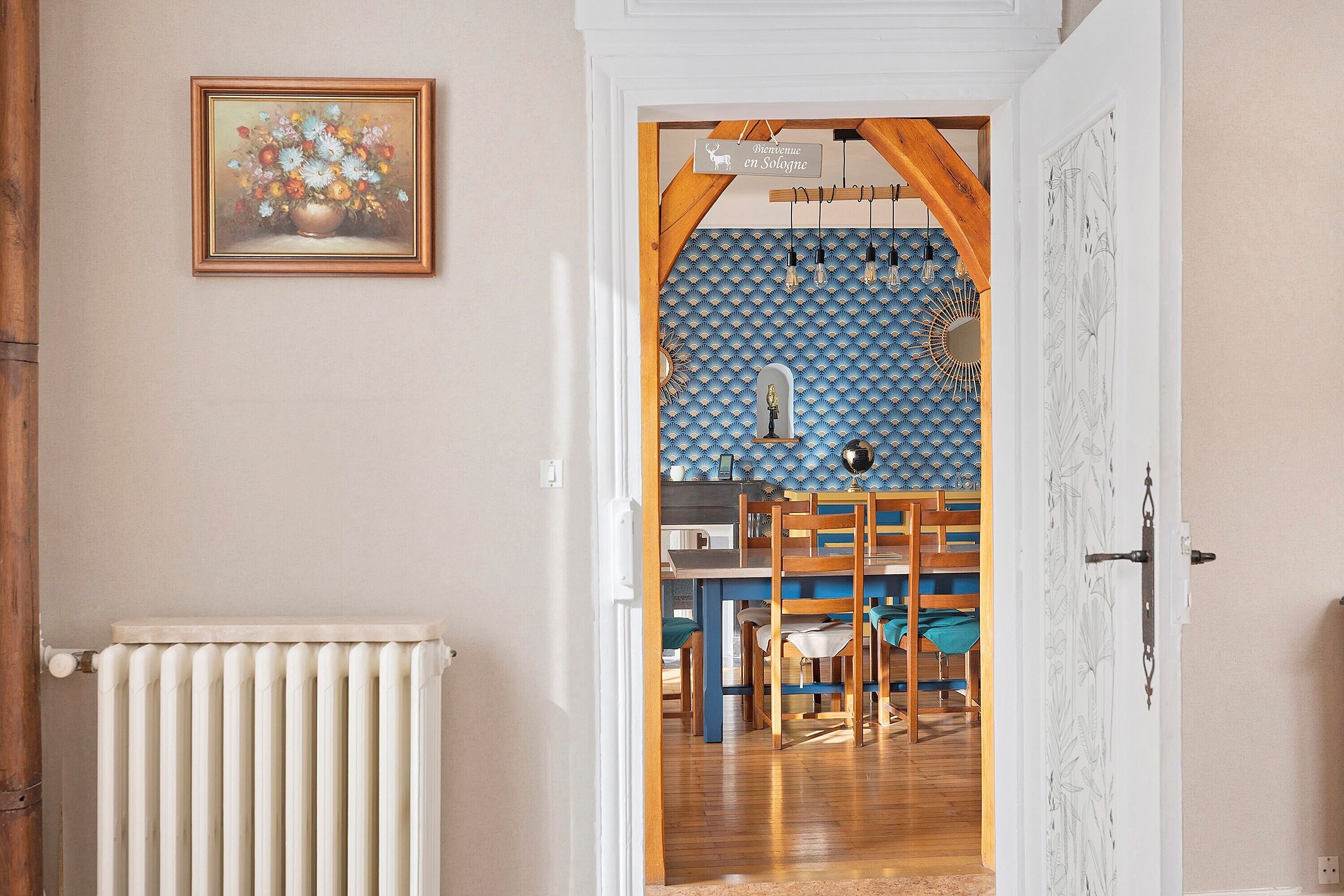 Une porte ouverte donne sur un espace repas avec un mur orné d’un motif bleu, un tableau floral encadré et un radiateur.