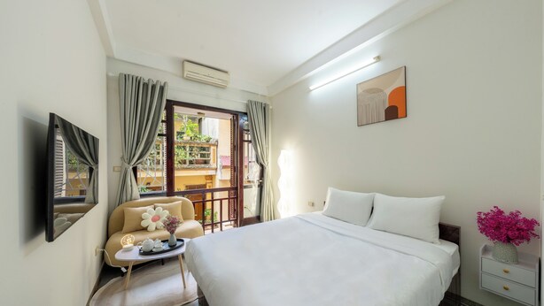 A hotel room with a bed, a small table with a vase of flowers, a chair, and a view of the outside balcony. - Homestay PW Hoang Sam (Hanoi)