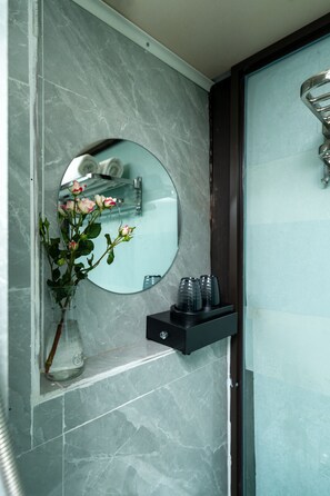 A bathroom with a marble wall, a round mirror, a vase with flowers, and a shelf with toiletries.